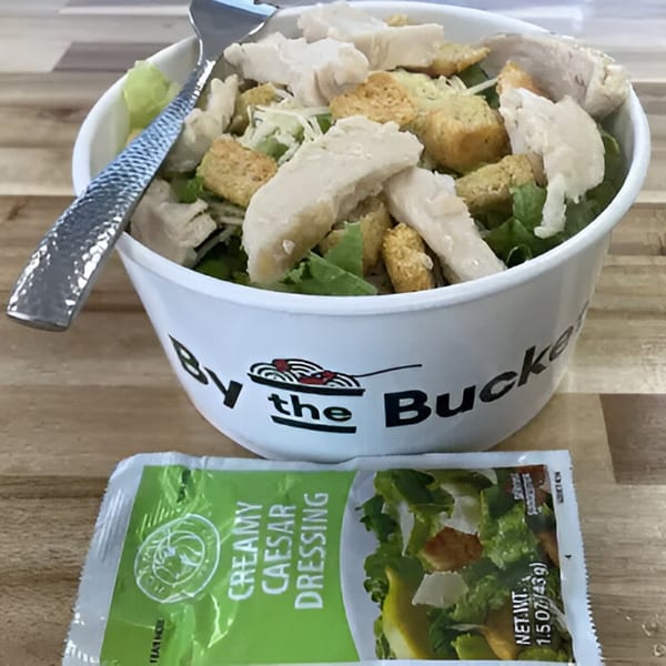 By The Bucket Caesar salad with chicken and croutons, alongside a packet of creamy Caesar dressing.
